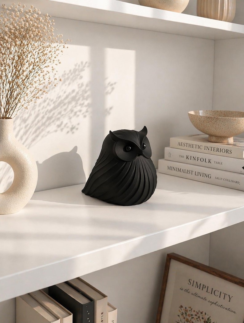 May include: A black owl figurine with detailed feather-like textures sits on a white shelf. The owl has large, round eyes and a slightly curved beak. The shelf also holds books, a vase with dried flowers, and a decorative bowl.