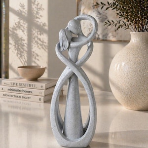 May include: A grey sculpture of two figures embracing, with intertwined arms and bodies. The abstract design features a kissing couple, with a textured surface. The sculpture is on a white surface, with books and a vase in the background.