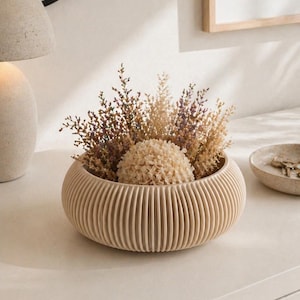 May include: A beige, ribbed, round decorative bowl filled with dried flowers and a spherical accent. The arrangement sits on a white surface, with a mirror and framed artwork in the background. The bowl's diameter is approximately 20 cm.