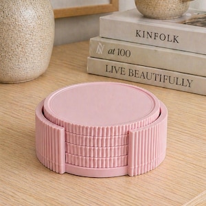 May include: A stack of four pink coasters with a ribbed edge design. The coasters are stacked on a light wooden surface. A vase with greenery, a mug, and books are in the background.