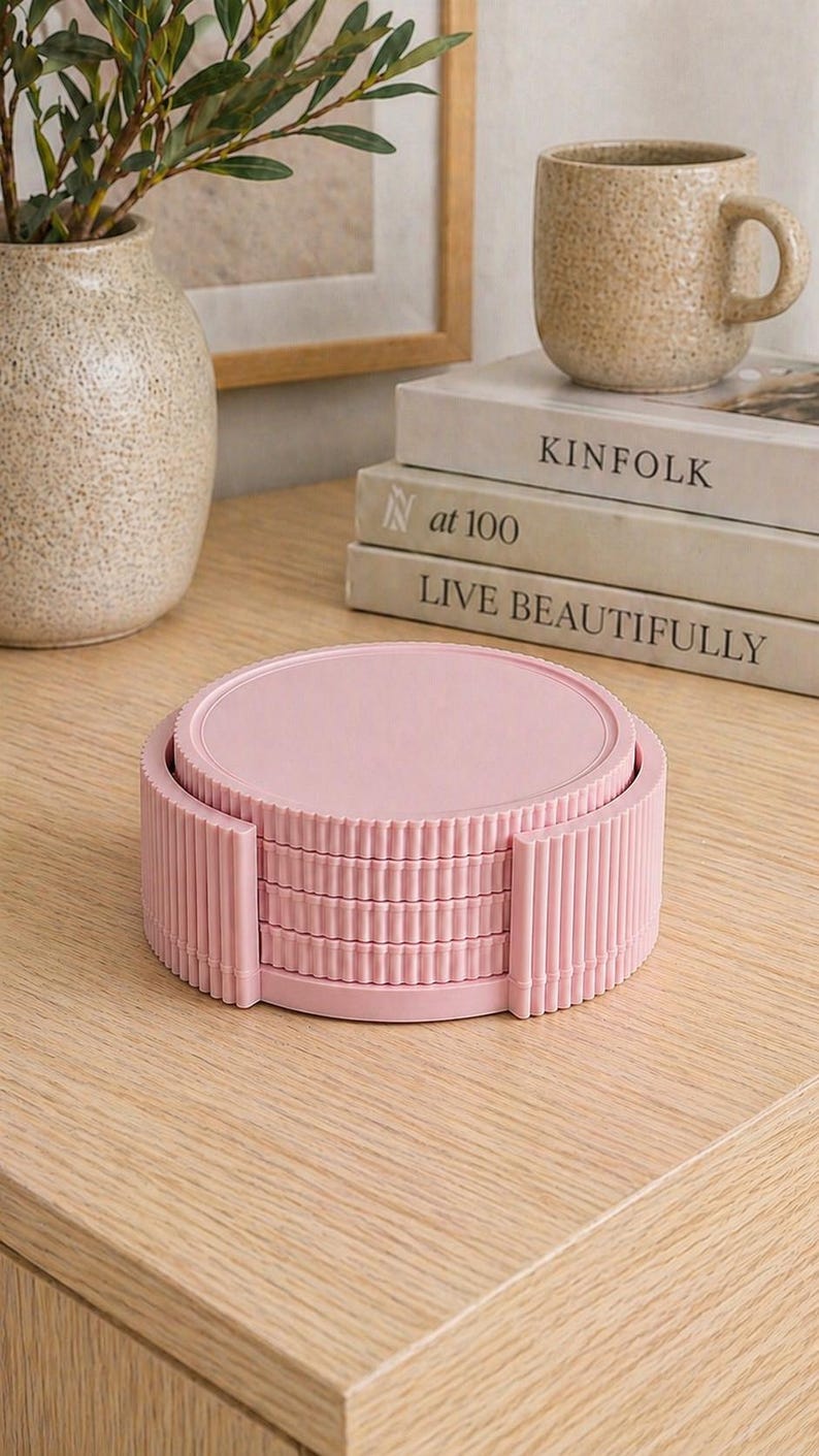May include: A stack of four pink coasters with a ribbed edge design. The coasters are stacked on a light wooden surface. A vase with greenery, a mug, and books are in the background.