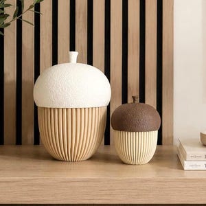 May include: Two acorn-shaped containers with removable lids. The larger container features a textured white cap and a light beige ribbed base. The smaller container has a dark brown textured cap and a light beige ribbed base. Both items are rounded.