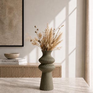 May include: An olive green, ribbed ceramic vase with a bulbous shape, filled with dried beige and brown flowers and grasses. The vase sits on a light-colored surface, with a neutral-toned background featuring a framed artwork and a wooden cabinet.