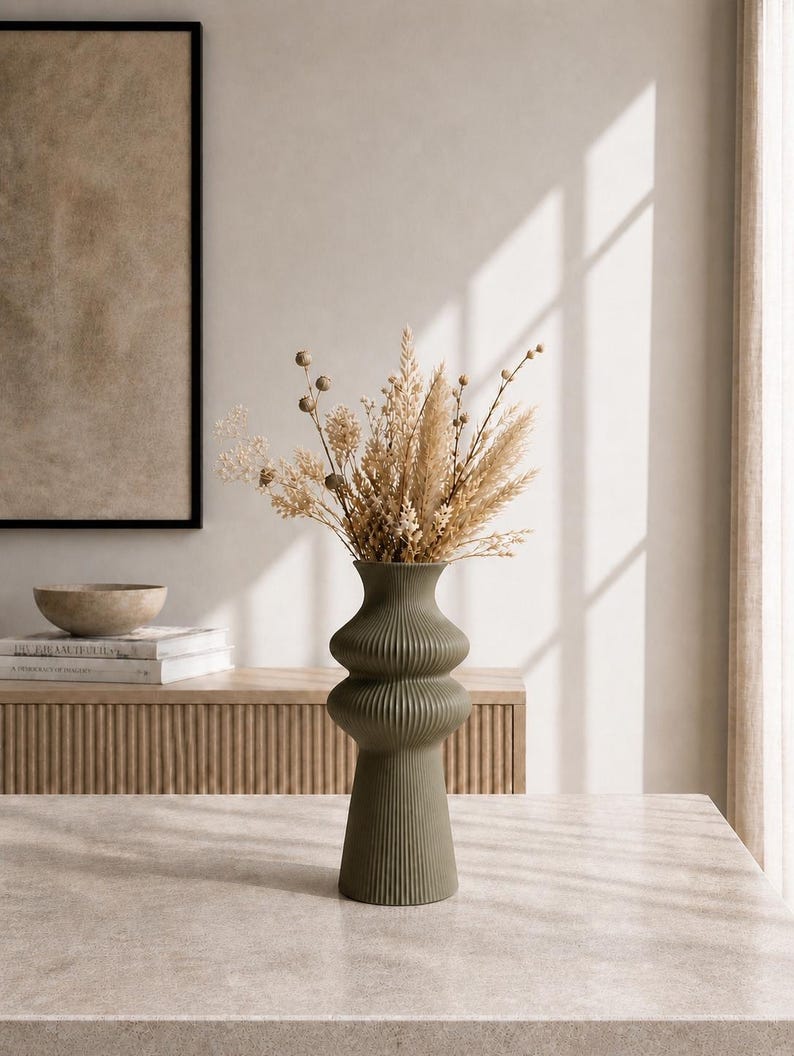 May include: An olive green, ribbed ceramic vase with a bulbous shape, filled with dried beige and brown flowers and grasses. The vase sits on a light-colored surface, with a neutral-toned background featuring a framed artwork and a wooden cabinet.