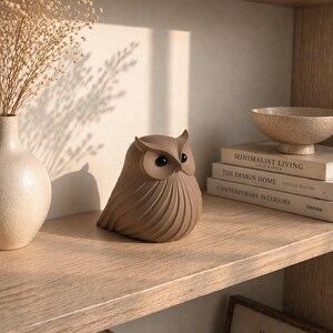 May include: A brown owl figurine with large eyes sits on a wooden shelf. The owl has a textured, ridged body. A vase with dried flowers and books are also on the shelf. The framed text reads "SIMPLE & SOPHISTICATED."