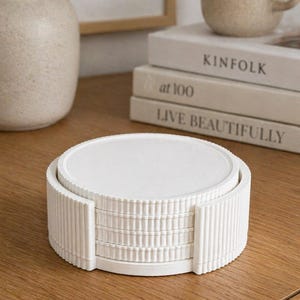 May include: A stack of white, round coasters with a ribbed edge, held in a matching holder. The coasters sit on a wooden surface, with a vase, mug, and books in the background.