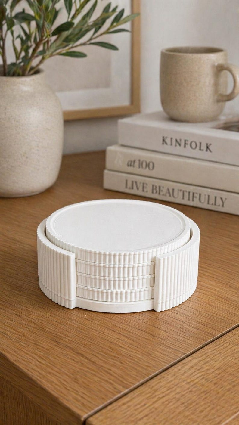 May include: A stack of white, round coasters with a ribbed edge, held in a matching holder. The coasters sit on a wooden surface, with a vase, mug, and books in the background.
