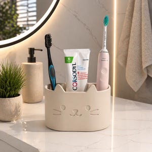May include: A beige cat-shaped toothbrush holder with a black and blue toothbrush, a tube of Corsodyl toothpaste, and a pink electric toothbrush. The holder sits on a white countertop in a bathroom setting.