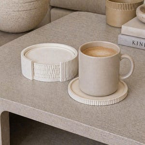 May include: A beige ceramic mug with a saucer, filled with a warm beverage, sits on a light gray table. A set of matching coasters is nearby. The scene suggests a cozy, relaxing atmosphere, perfect for enjoying a hot drink.