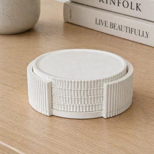 May include: A set of white, round coasters with a ribbed holder. The coasters are stacked neatly on a light-colored wooden surface. The image also shows a potted plant and two books with the titles "KINFOLK" and "LIVE BEAUTIFULLY".