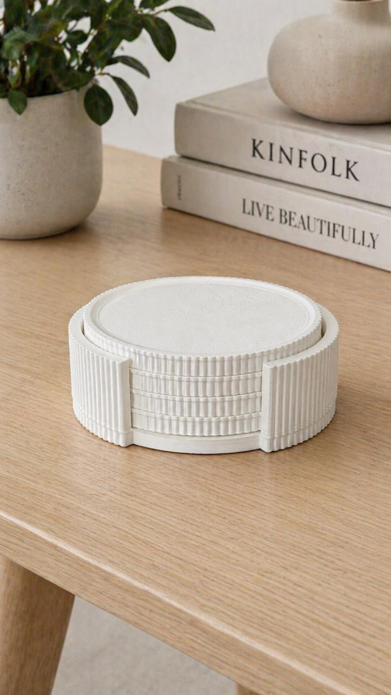 May include: A set of white, round coasters with a ribbed holder. The coasters are stacked neatly on a light-colored wooden surface. The image also shows a potted plant and two books with the titles "KINFOLK" and "LIVE BEAUTIFULLY".