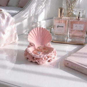 May include: A pink seashell-shaped jewelry dish holds two rings with gold bands and a diamond. The dish sits on a white surface, next to a pink book with the words "SELF LOVE" and two bottles of perfume.
