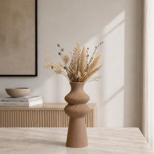 May include: A tall, brown, ribbed vase with a unique, stacked design, filled with dried flowers and grasses. The vase is on a light-colored table, with a neutral-toned background. The vase is approximately 12 inches tall.