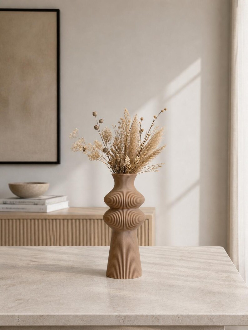 May include: A tall, brown, ribbed vase with a unique, stacked design, filled with dried flowers and grasses. The vase is on a light-colored table, with a neutral-toned background. The vase is approximately 12 inches tall.