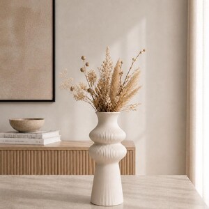 May include: A white, ribbed ceramic vase with a bulbous shape, filled with dried beige and tan grasses and flowers. The vase sits on a light-colored surface, with a wooden cabinet and framed artwork in the background. Home decor.