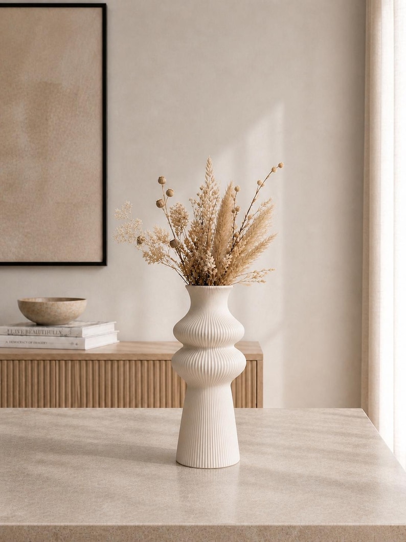 May include: A white, ribbed ceramic vase with a bulbous shape, filled with dried beige and tan grasses and flowers. The vase sits on a light-colored surface, with a wooden cabinet and framed artwork in the background. Home decor.