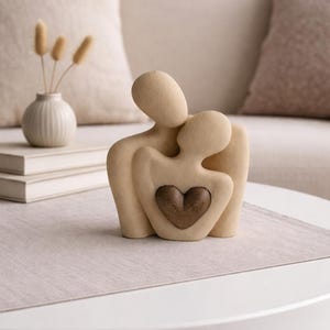 May include: A beige sculpture of two embracing figures with a heart-shaped cutout. The sculpture is on a white table with a beige runner. A small white vase with dried plants and a stack of books are in the background.