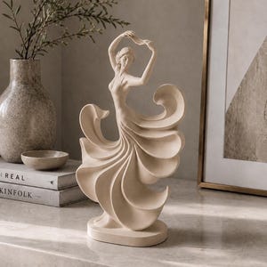 May include: A cream-coloured figurine of a dancer in a flowing dress with large, sweeping, fan-like folds. The figure stands on an oval base with arms raised above the head, capturing a dynamic pose with smooth, matte surfaces and curved lines.