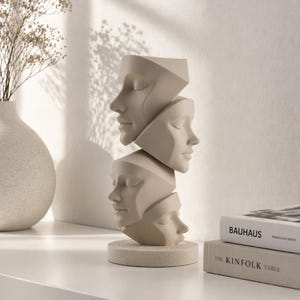 May include: A beige sculpture of four abstract faces stacked on top of each other. The faces have closed eyes and are set on a round base. Decorative books and a vase are in the background. The sculpture is a modern art piece.