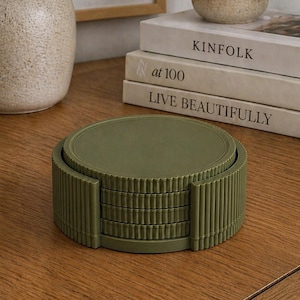 May include: A stack of olive green coasters with a ribbed edge design. The coasters are stacked on a wooden surface, next to a beige mug and a stack of books. A vase with greenery is in the background.