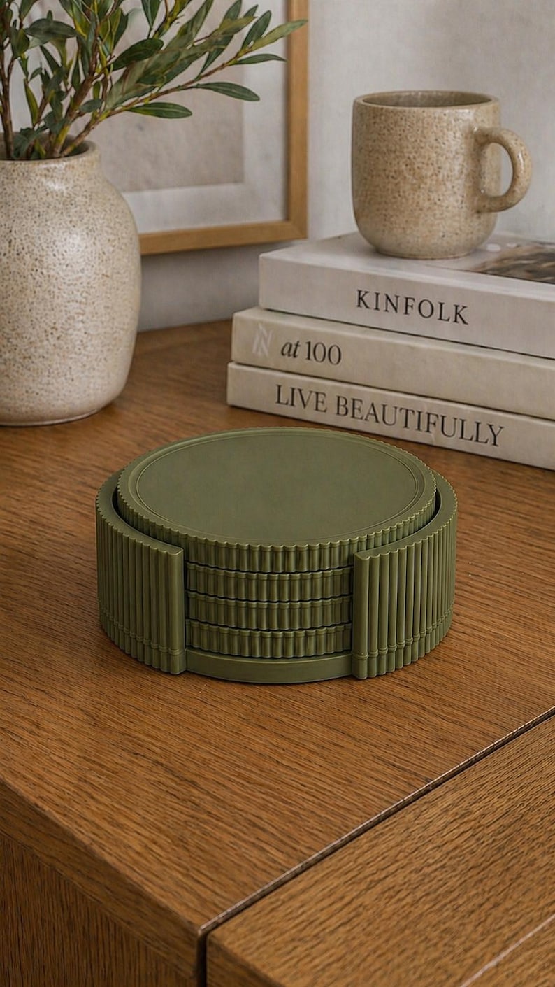 May include: A stack of olive green coasters with a ribbed edge design. The coasters are stacked on a wooden surface, next to a beige mug and a stack of books. A vase with greenery is in the background.