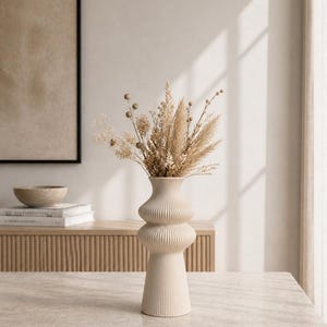 May include: A cream-colored, ribbed ceramic vase with a bulbous shape, filled with dried beige and tan flowers and grasses. The vase sits on a light-colored stone table. A wooden cabinet and framed artwork are in the background.