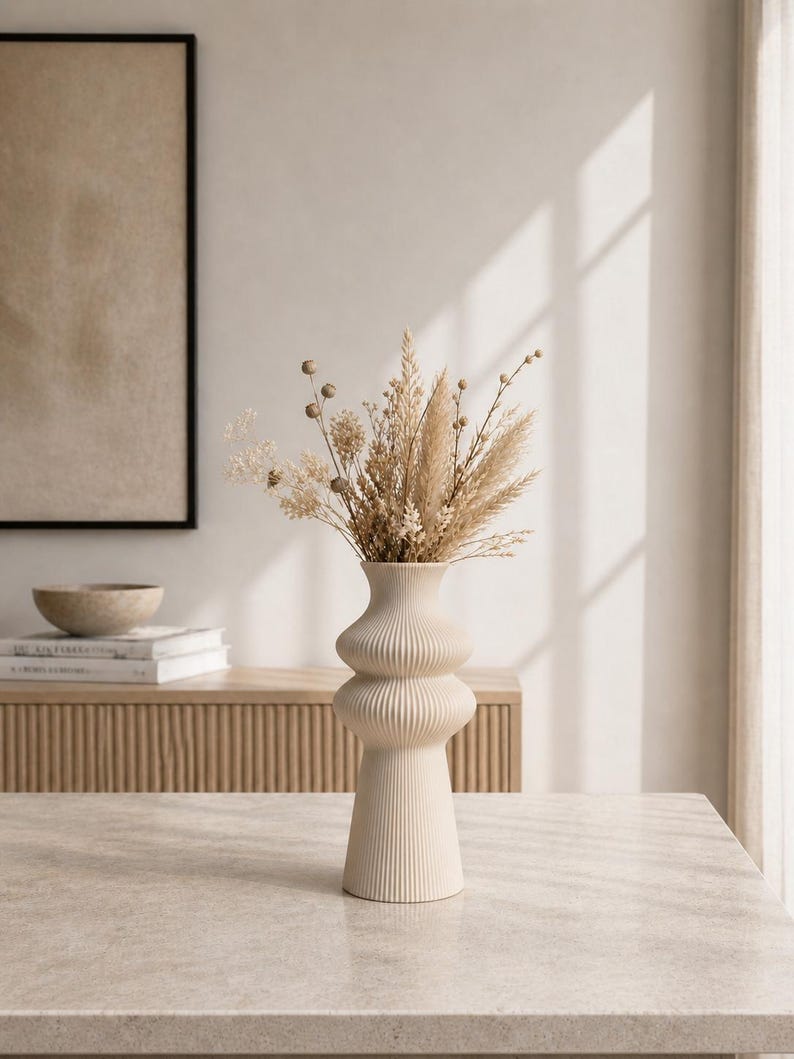 May include: A cream-colored, ribbed ceramic vase with a bulbous shape, filled with dried beige and tan flowers and grasses. The vase sits on a light-colored stone table. A wooden cabinet and framed artwork are in the background.