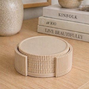 Può includere: Un set di sottobicchieri beige con bordo a coste, su una superficie in legno chiaro. Sullo sfondo, una tazza beige abbinata e dei libri. È visibile anche un vaso con della vegetazione.