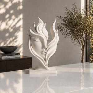 May include: A white sculpture of a serene face emerging from stylized flames. The artwork is set on a white rectangular base, with soft shadows and natural light enhancing its form. The sculpture is a decorative piece.