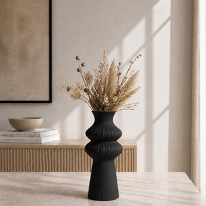 May include: A black, ribbed ceramic vase with a unique hourglass shape, filled with dried beige and brown flowers and grasses. The vase sits on a light-colored stone table, with a neutral-toned background.
