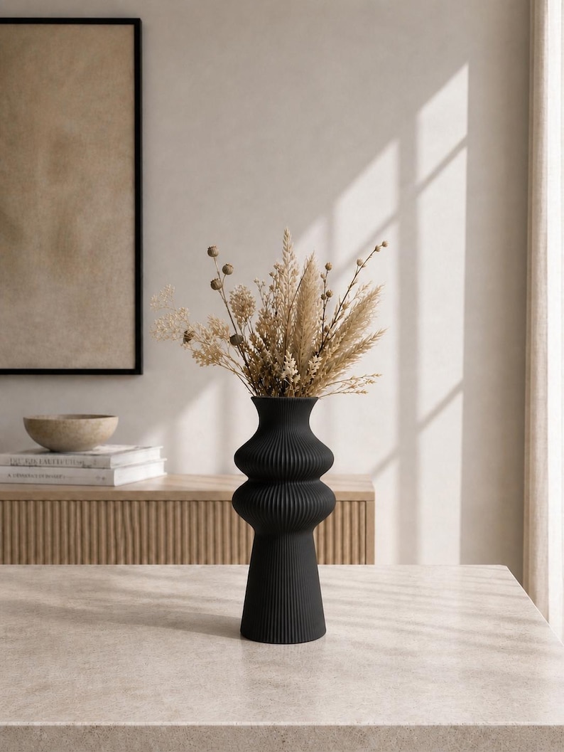 May include: A black, ribbed ceramic vase with a unique hourglass shape, filled with dried beige and brown flowers and grasses. The vase sits on a light-colored stone table, with a neutral-toned background.
