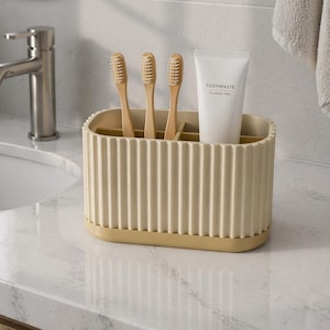 Puede incluir: Un soporte para cepillos de dientes beige con tres cepillos de dientes de madera y un tubo de pasta de dientes. El soporte tiene un diseño acanalado y se asienta sobre una base amarillo claro. El tubo de pasta de dientes es blanco con texto negro.
