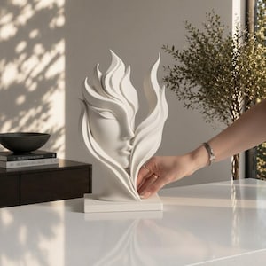 May include: A white sculpture of a face with flowing, flame-like shapes. The sculpture is on a white base and sits on a white surface. The sculpture is a decorative item.
