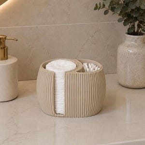 Könnte beinhalten: Ein beigefarbener, gerippter Organizer mit zwei Fächern für Wattepads und Wattestäbchen. Der Organizer steht auf einer Arbeitsplatte, neben einem Seifenspender und einer Vase mit Grünzeug. Er ist mit weißen Wattepads und Wattestäbchen gefüllt.