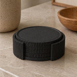 May include: A set of six black coasters with a ridged, vertical design on the sides. The coasters are stacked in a holder, and the top coaster is visible. The coasters are made of a matte black material and are designed to protect surfaces from drink rings.