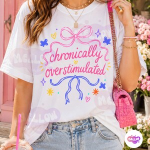 May include: A white short-sleeve t-shirt featuring a pink bow at the top and a blue bow at the bottom, with the pink text "chronically overstimulated" centered between them, surrounded by small colorful stars, hearts, and butterfly graphics.