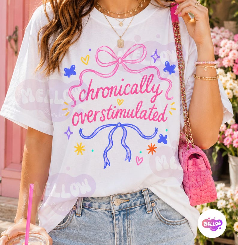 May include: A white short-sleeve t-shirt featuring a pink bow at the top and a blue bow at the bottom, with the pink text "chronically overstimulated" centered between them, surrounded by small colorful stars, hearts, and butterfly graphics.