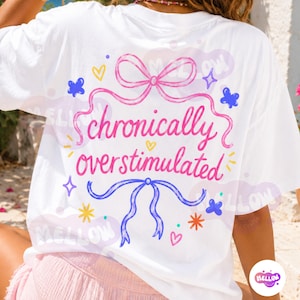 May include: A white t-shirt featuring a colorful graphic on the back with the text "chronically overstimulated" in pink cursive. The design includes a pink bow at the top, a blue bow at the bottom, and various small yellow, blue, and orange icons.