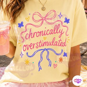 May include: A pale yellow t-shirt featuring the pink text "chronically overstimulated" centered between a pink bow at the top and a blue bow at the bottom. The design includes small blue butterflies, yellow hearts, and colorful starburst accents.