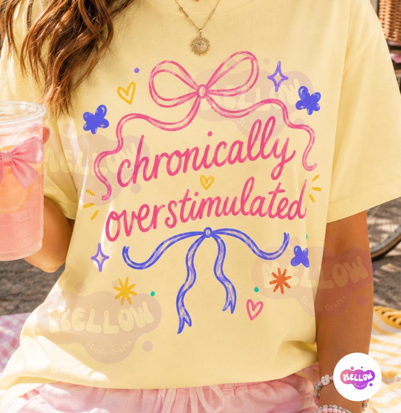 May include: A pale yellow t-shirt featuring the pink text "chronically overstimulated" centered between a pink bow at the top and a blue bow at the bottom. The design includes small blue butterflies, yellow hearts, and colorful starburst accents.