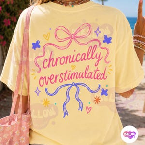 May include: A pale yellow t-shirt featuring a pink ribbon graphic at the top and a blue ribbon graphic at the bottom. The text "chronically overstimulated" is printed in pink cursive in the center, surrounded by small blue and yellow stars.