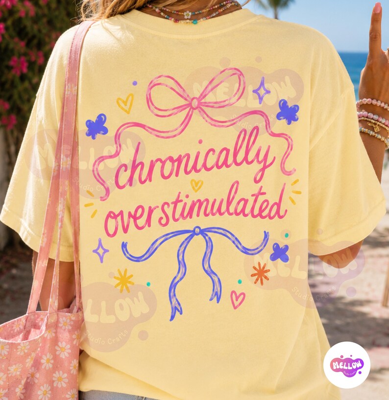 May include: A pale yellow t-shirt featuring a pink ribbon graphic at the top and a blue ribbon graphic at the bottom. The text "chronically overstimulated" is printed in pink cursive in the center, surrounded by small blue and yellow stars.