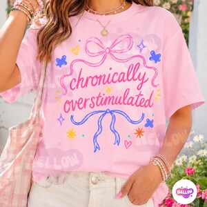May include: A light pink t-shirt featuring the text "chronically overstimulated" in a dark pink script font. The design includes a pink bow at the top, a blue bow at the bottom, and various small colorful hearts, stars, and butterfly graphics.