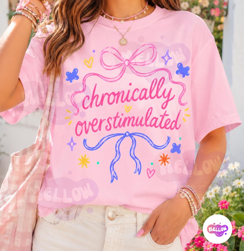 May include: A light pink t-shirt featuring the text "chronically overstimulated" in a dark pink script font. The design includes a pink bow at the top, a blue bow at the bottom, and various small colorful hearts, stars, and butterfly graphics.