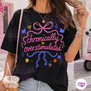May include: A black short-sleeved t-shirt featuring a graphic print with the text "chronically overstimulated" in pink cursive lettering. The design includes pink and blue ribbon bows, small yellow and orange stars, and tiny hearts.