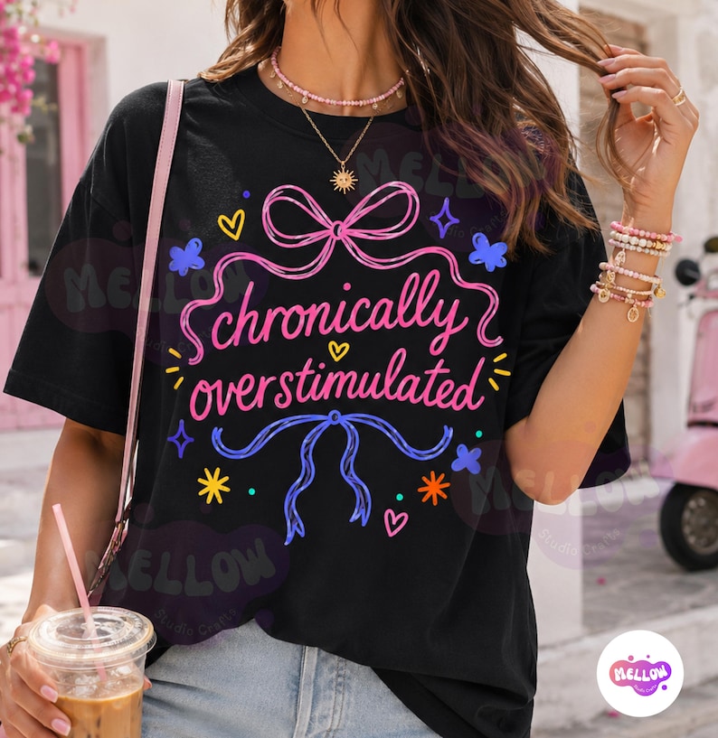 May include: A black short-sleeved t-shirt featuring a graphic print with the text "chronically overstimulated" in pink cursive lettering. The design includes pink and blue ribbon bows, small yellow and orange stars, and tiny hearts.