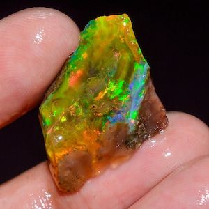 May include: A raw, unpolished opal gemstone displaying vibrant flashes of neon green, bright orange, and deep blue play-of-colour across its surface. The stone features a translucent, irregular shape with a textured, earthy brown base.