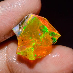 May include: A raw, unpolished opal gemstone displaying vibrant flashes of neon orange, bright yellow, and electric green play-of-color. The stone has an irregular, jagged shape with a translucent, glassy texture and a multifaceted surface.