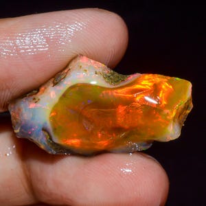 Peut inclure: Une opale brute non polie présentant un jeu de couleurs vibrant avec des éclats intenses d'orange, de rouge et de jaune sur une base translucide blanc laiteux. La pierre a une forme organique irrégulière avec des inclusions rocheuses visibles.