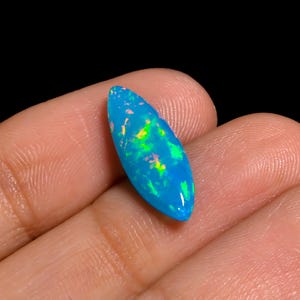 May include: A marquise-cut opal gemstone featuring a vibrant blue base color with internal flashes of bright green and orange light. The stone has a smooth, polished surface and a pointed, elongated oval shape with a domed top profile.